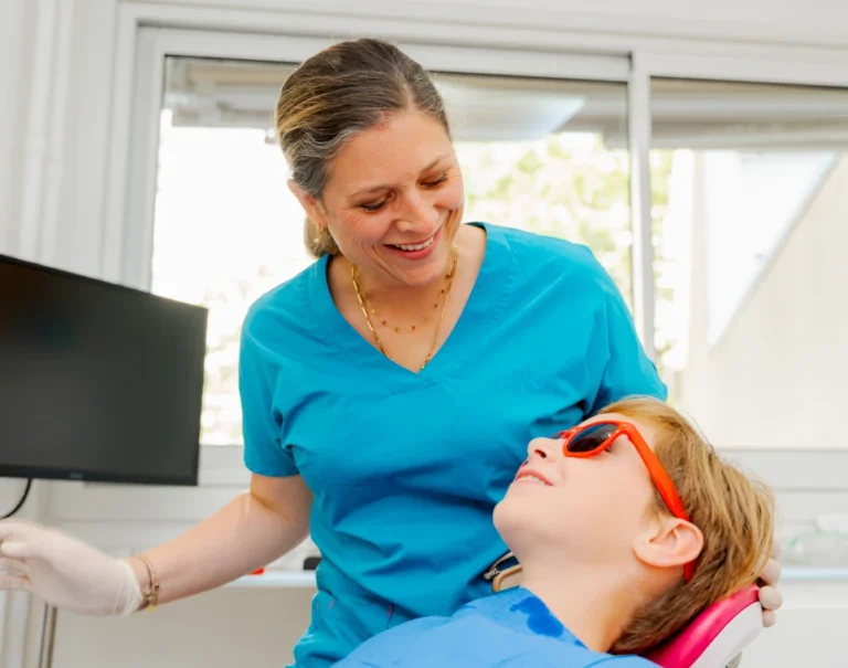 Dentiste souriante auprès d’un enfant allongé au fauteuil, lunettes de protection